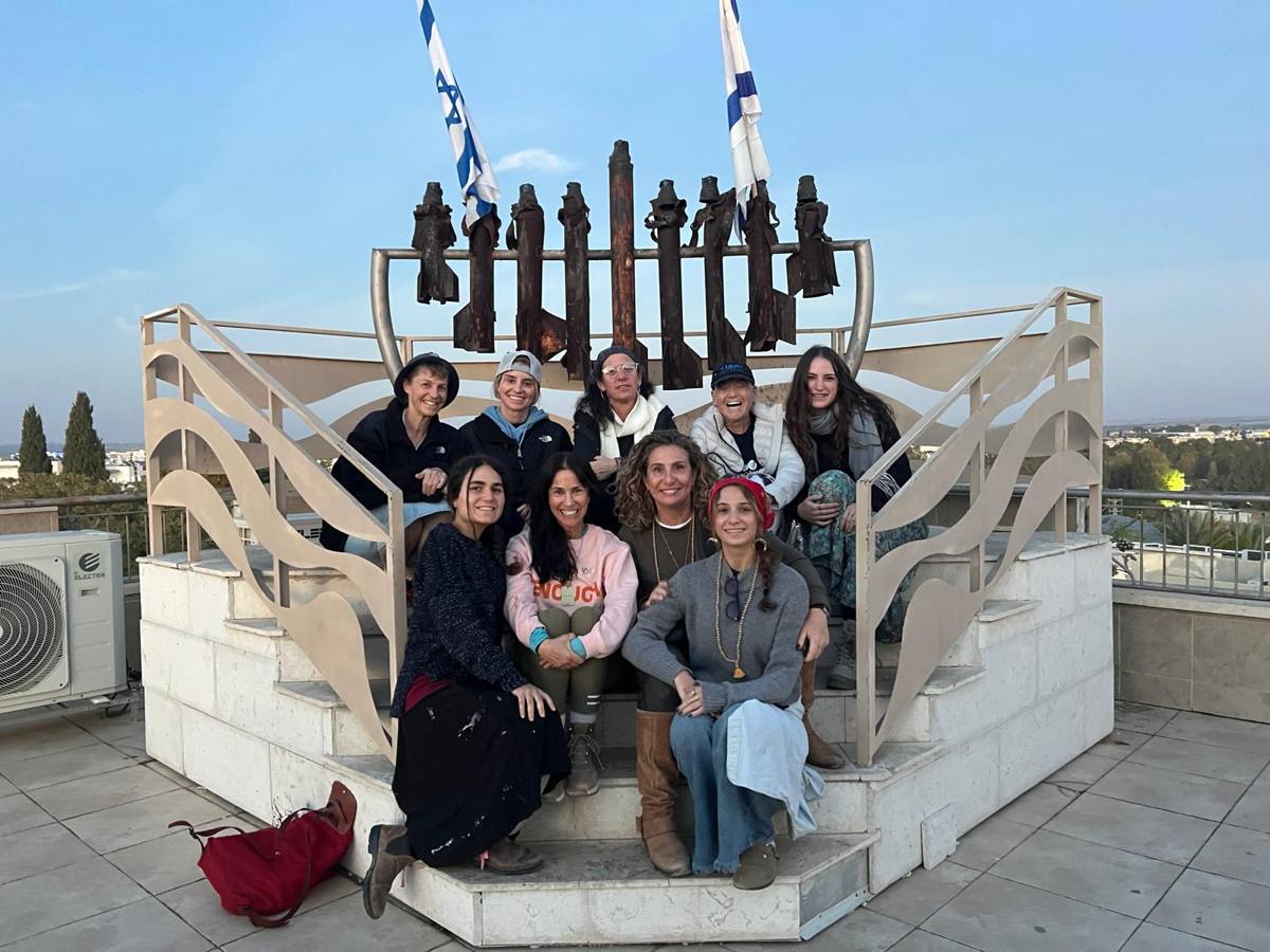 The six women on the mission, with three friends, sitting on the steps beneath a large iron menorah in Israel, two Israeli flags behind them